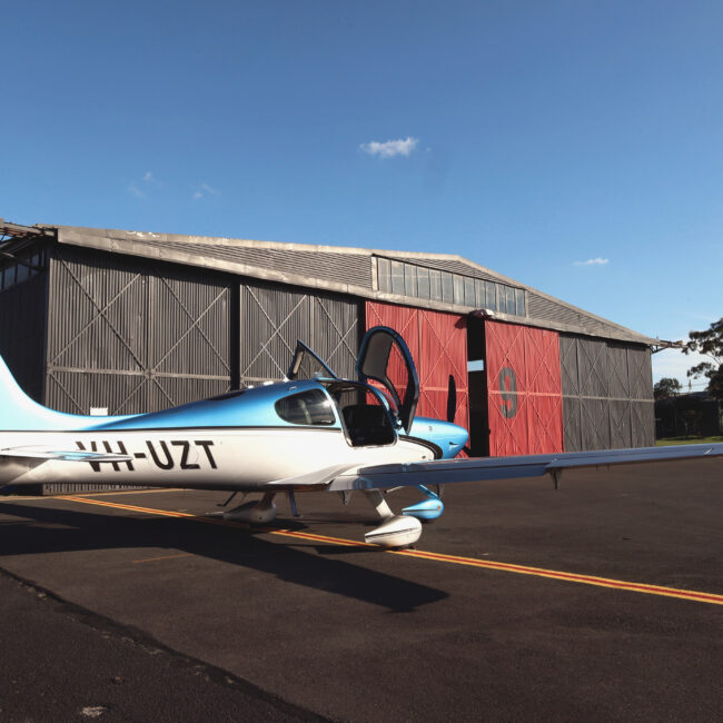 Cirrus SR20 aircraft parked outside Avia Aviation hangar under a blue sky with scattered clouds.