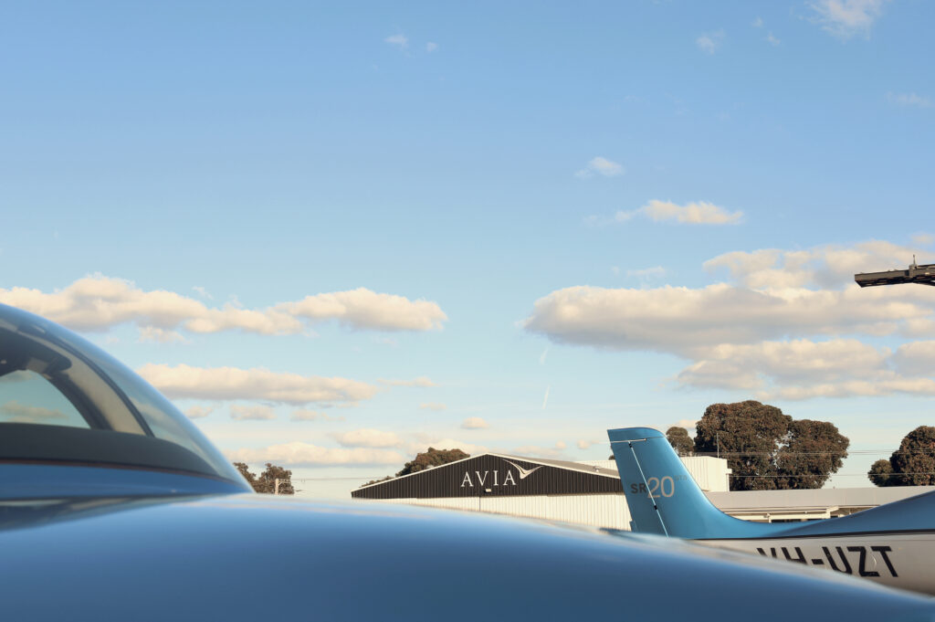 Cirrus SR20 aircraft parked at Avia Aviation hangar under a clear blue sky with scattered clouds.