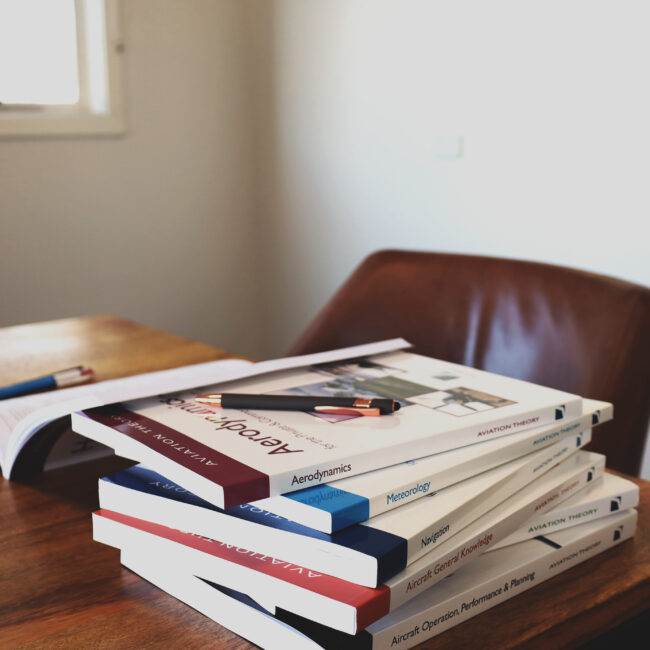 Stack of aviation theory books on a wooden desk, including titles on aerodynamics, navigation, and meteorology, used for pilot training study.