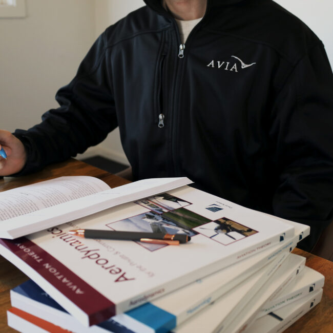 Student pilot studying aviation theory books at a desk, wearing an Avia Aviation jacket, preparing for ground school training.