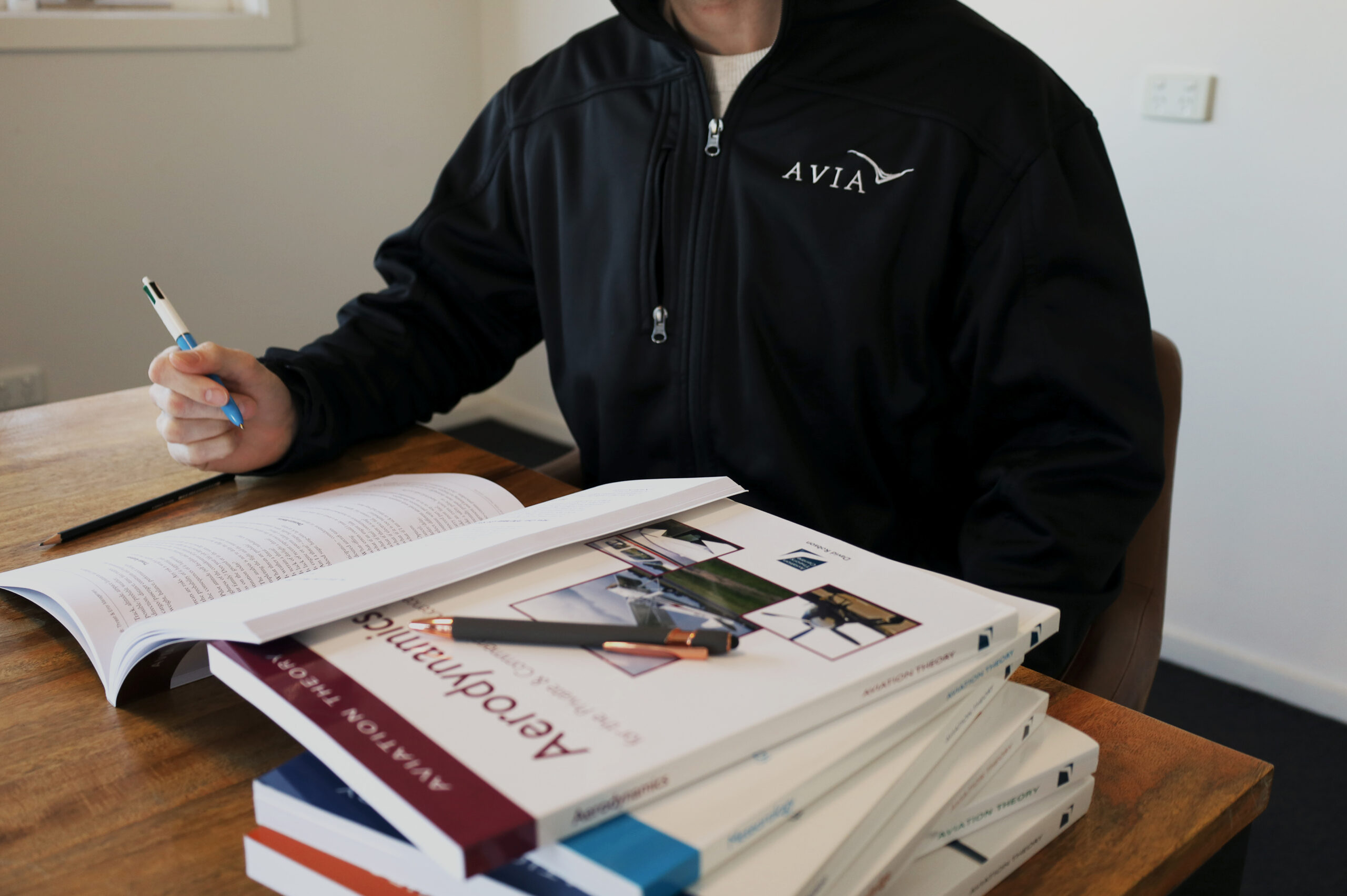 Student pilot studying aviation theory books at a desk, wearing an Avia Aviation jacket, preparing for ground school training.