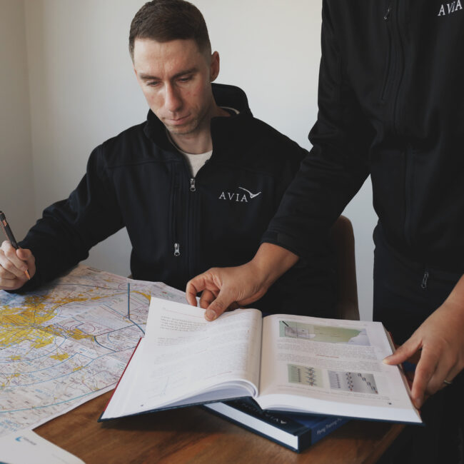 Student pilots in Avia Aviation uniforms reviewing flight planning charts and medical certification requirements during ground school preparation.