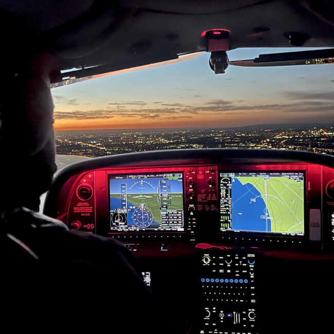 Pilot flying at night with illuminated cockpit displays showing navigation systems and city lights visible through the windshield at sunset.
