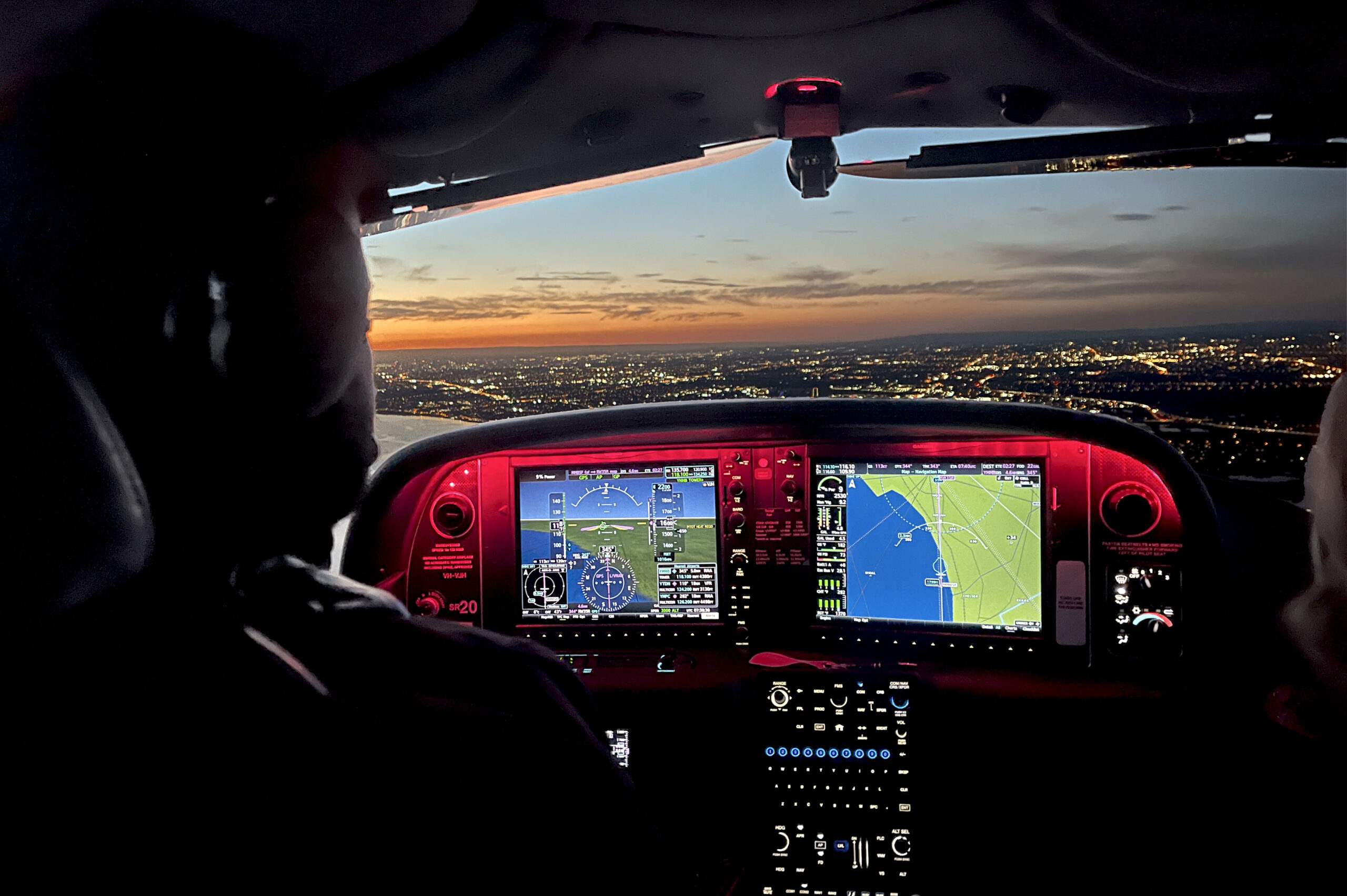 Pilot flying at night with illuminated cockpit displays showing navigation systems and city lights visible through the windshield at sunset.