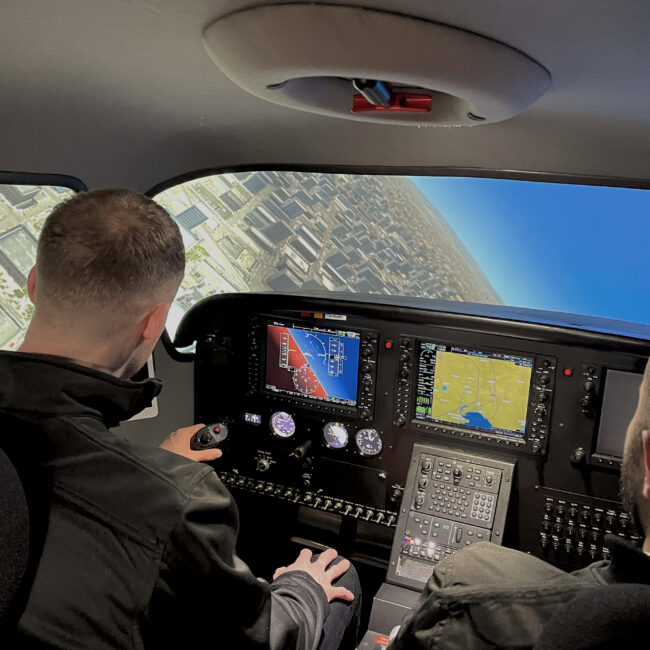 Student pilot practicing a turn in a full-motion flight simulator under instructor supervision, with cityscape visible on the screen.