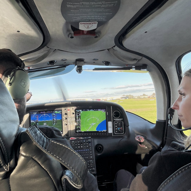 Flight instructor and student pilot seated in a Cirrus SR20 cockpit, reviewing instruments during a pre-takeoff briefing on a clear day.