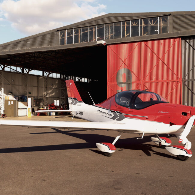 Red and white P-Mentor training aircraft parked on the tarmac in front of an open hangar at sunset.