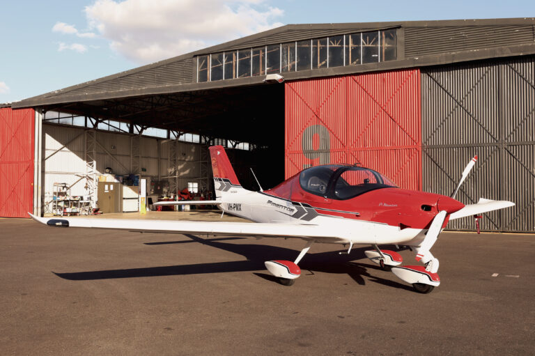 Red and white P-Mentor training aircraft parked on the tarmac in front of an open hangar at sunset.