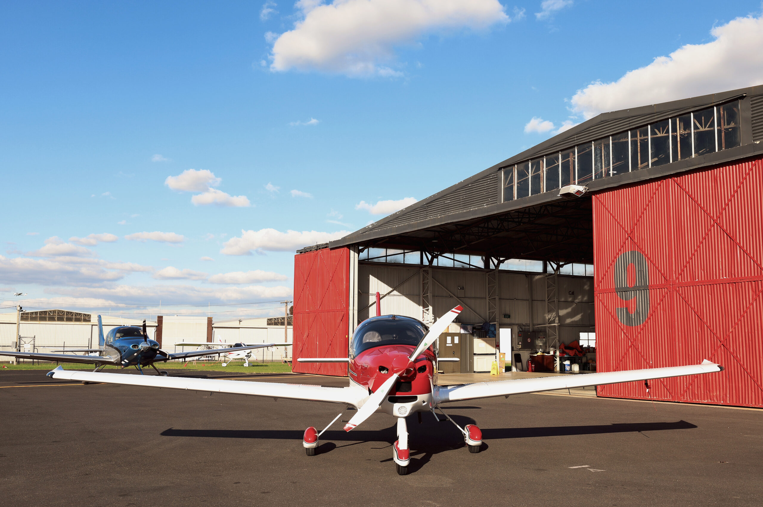 Red and white training aircraft parked outside Hangar 9 at Moorabbin Airport, representing Avia Aviation's facilities for overseas pilot licence conversion in Australia.