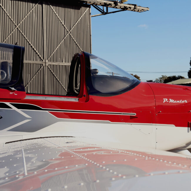 Side view of a red and white P-Mentor training aircraft parked on the tarmac in front of a hangar, used for student flight lessons at Avia Aviation.